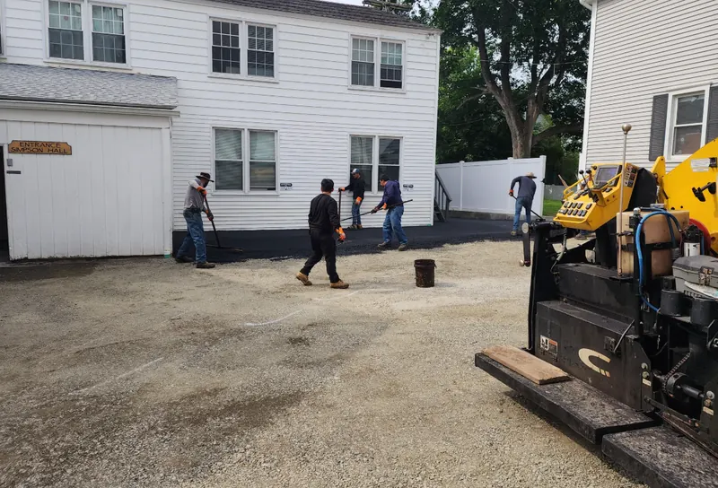 A paving crew raking hot asphalt behind a paver machine in a parking area beside a white clapboard building.