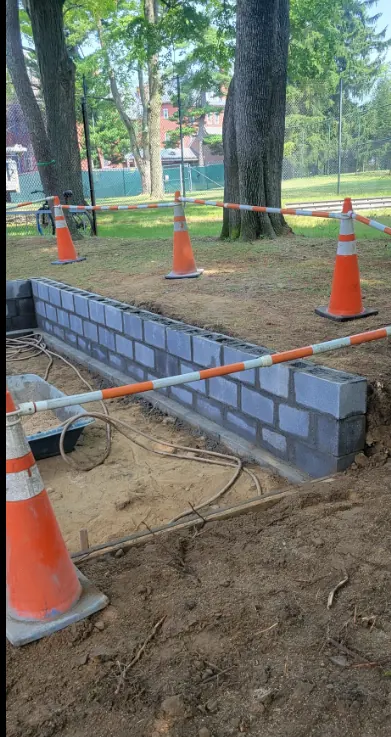 The first courses of a concrete block wall laid to line in an excavated trench, with safety cones and tape around the work.