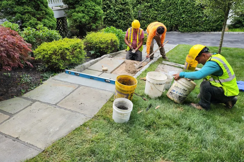 Three crew members in hard hats and hi-vis setting large bluestone slabs into a screeded bed beside a lawn.