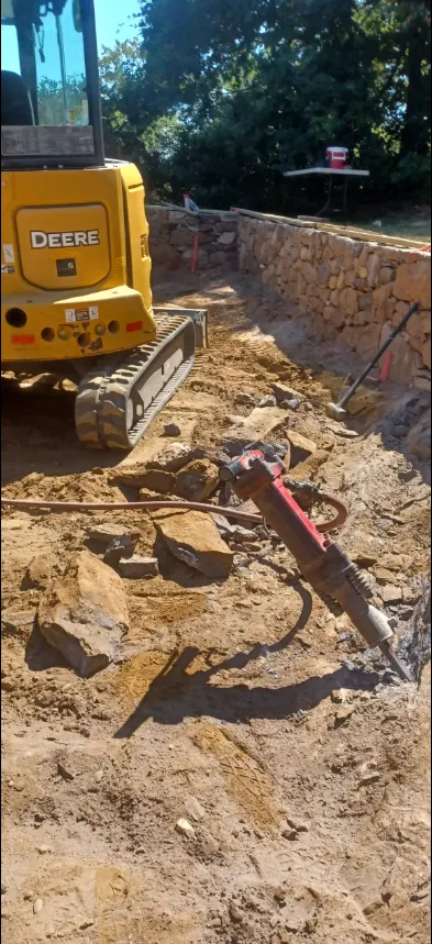 A mini excavator and a hydraulic breaker working on rock in an open trench alongside a completed dry-laid stone wall.