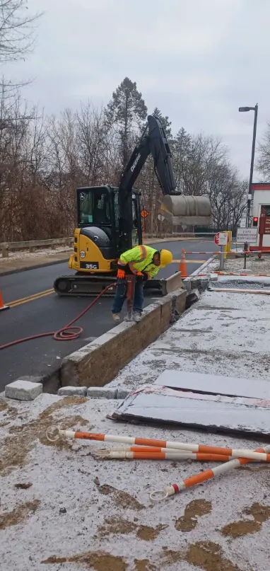 A worker with a breaker cutting out a failed concrete sidewalk section beside a road, with an excavator and new slabs staged nearby.