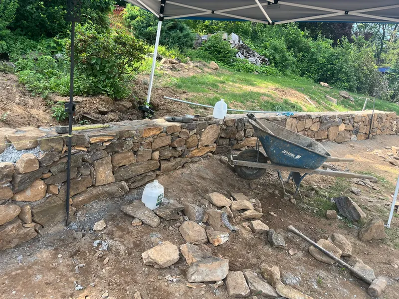 A dry-laid fieldstone retaining wall part-built under a shade canopy, with a wheelbarrow and loose stone in the foreground.