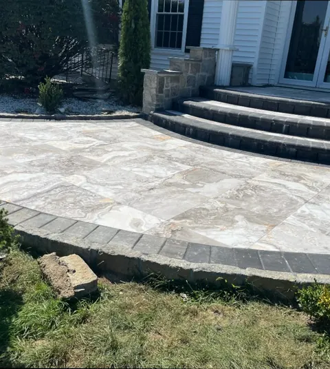 A pale porcelain paver patio with a charcoal border, curving up to paver steps and a stone pier at a house entrance.
