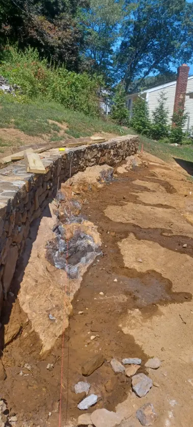 A stone retaining wall cut into a grassed slope beside a house, with the trench behind it open and drainage stone going in.