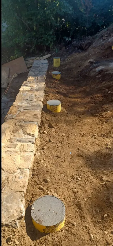 A natural stone retaining wall under construction, with poured concrete pier footings visible in the excavated trench beside it.